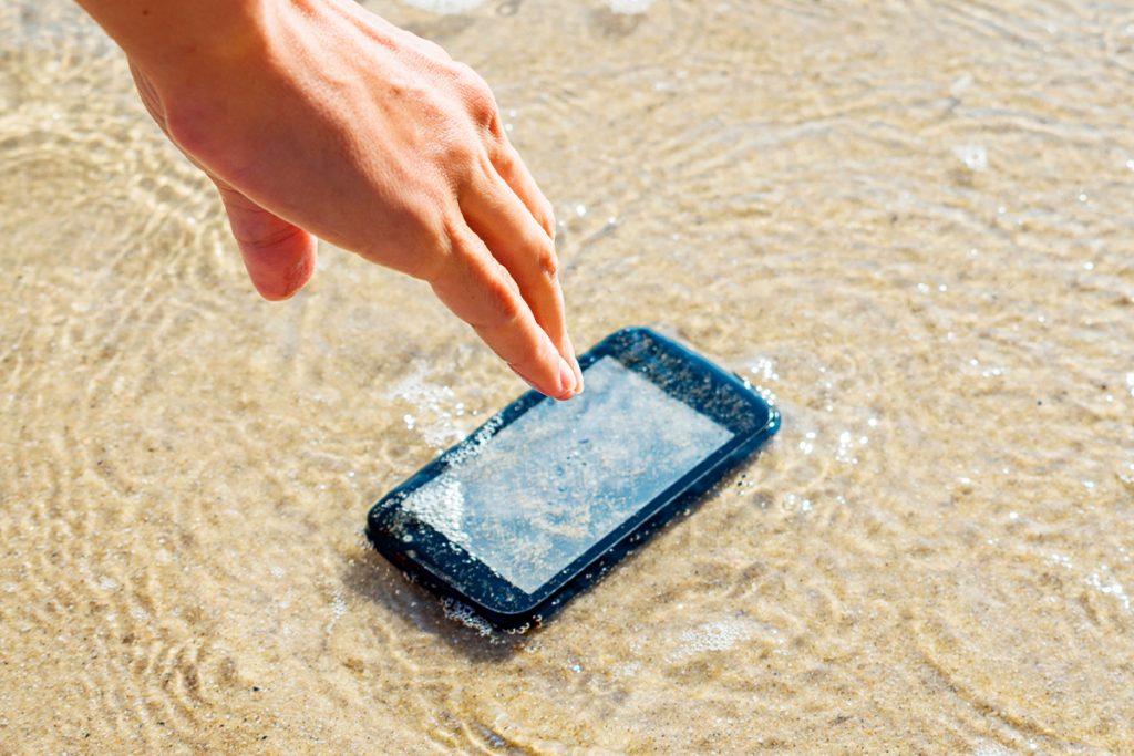 Capa blog-post celular caiu na água. Aparelho na areia, coberto pela água no mar, com uma mão tentando alcançá-lo.