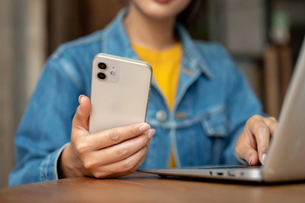 Pessoa segurando um iPhone e usando um MacBook, ilustrando como verificar se seu iPhone é desbloqueado.