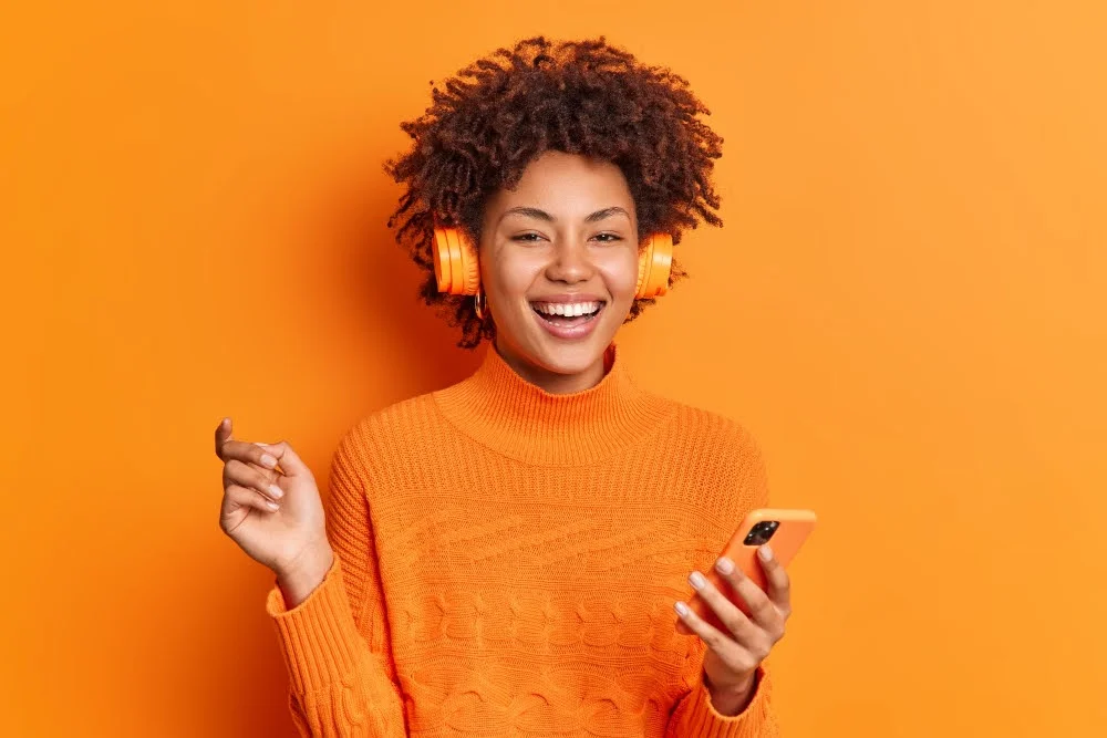 mulher sorrindo com suéter laranja, fones de ouvido e celular na mão em fundo laranja