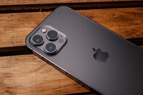 iPhone 12 Pro Max na cor cinza chumbo. Aparelho está virado para baixo em cima de uma mesa de madeira. 
