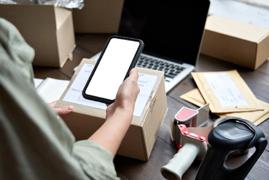 Mulher segurando um smartphone perto de uma mesa. Na mesa, vemos uma caixa para embalagem de transporte do celular, como se ele fosse ser embalado para entrega.
