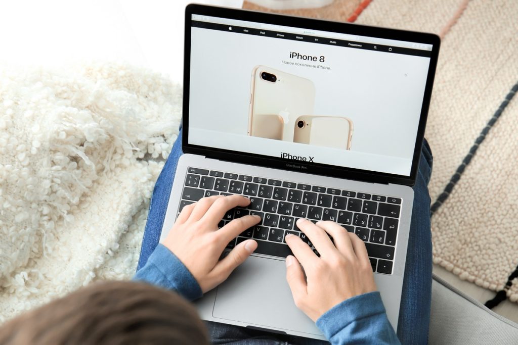Homem com as duas mãos sobre o teclado de um MacBook. Na tela do computador, vemos um anúncio de um iPhone 8.