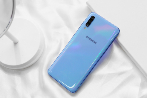 Samsung Galaxy A70 azul claro sobre uma mesa com toalha branca. O telefone está com a tela virada para baixo. 