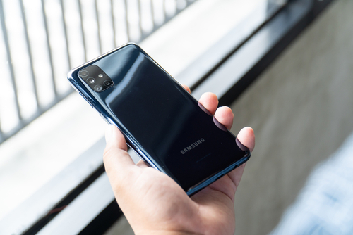  Mão de uma pessoa segurando um celular Samsung Galaxy M51 preto celeste. O telefone está com a tela virada para baixo. 