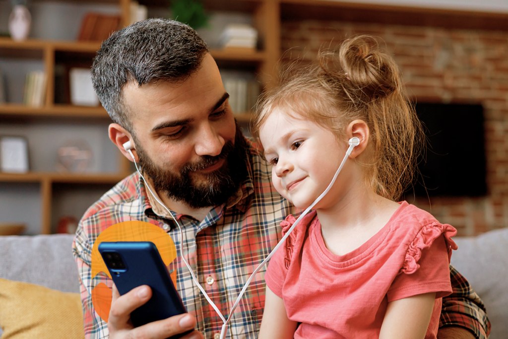 Foto de pai e filha olhando um smartphone que está nas mãos do homem. A criança está sentada no coloco do pai, com fones de ouvido na orelha.