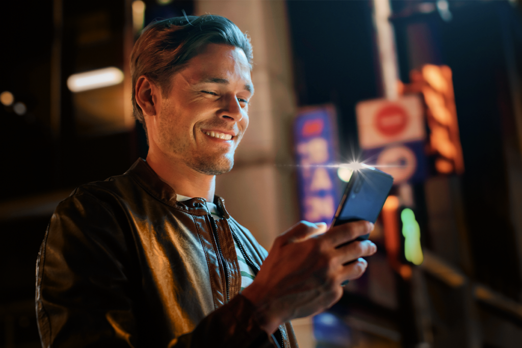 Homem em primeiro plano, com um celular na mão direita, sorrindo para a tela do aparelho.