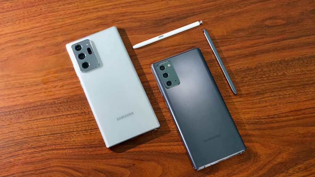 Dois celulares sobre uma mesa de madeira, ambos com a tela virada para baixo. Na esquerda, um Note 20 cinza claro, acompanhado de caneta S Pen. À direita, um Note 20 Ultra cinza chumbo, acompanhado de caneta S Pen, da mesma cor.