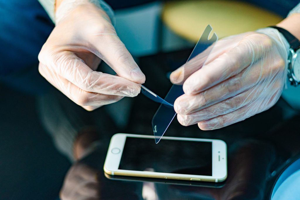 Pessoa com luvas cirúrgicas nas mãos aplicando uma película de vidro em um iPhone.