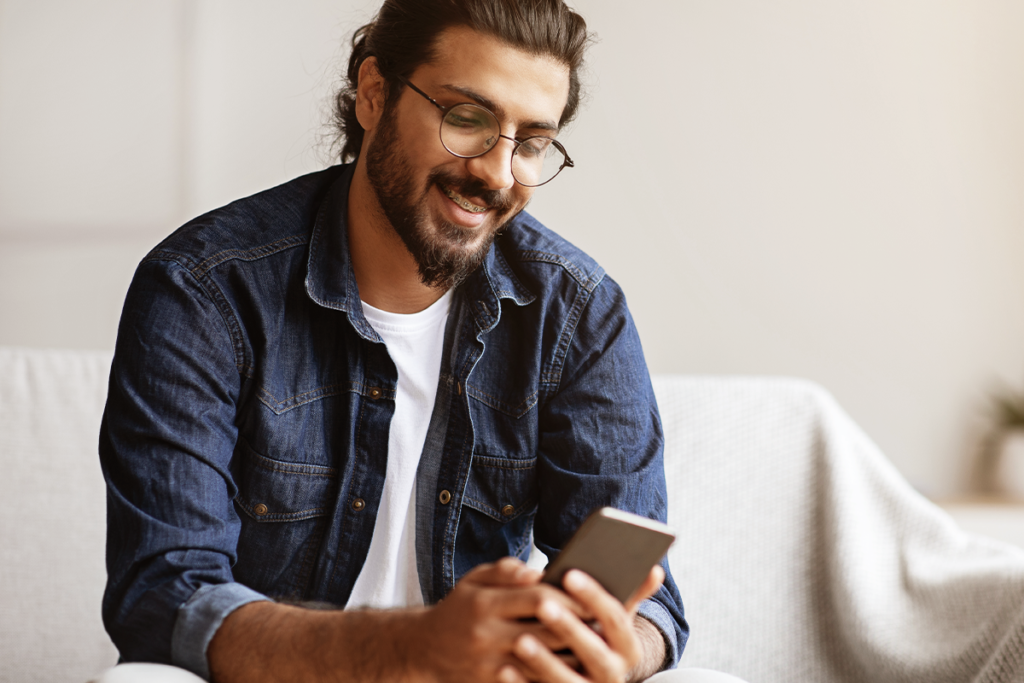 Homem com blusa jeans sentado em um sofá branco, com um celular nas mãos. Ele está sorrindo, olhando para a tela do aparelho.