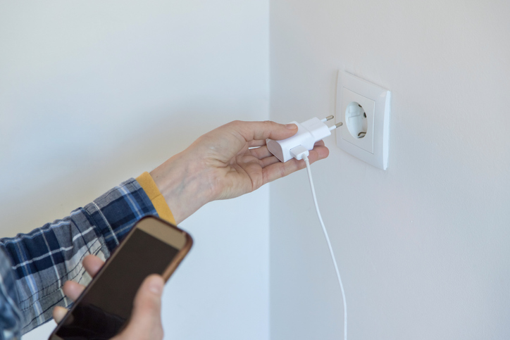 Mão esquerda masculina colocando um carregador branco em uma tomada de parede branca. Na mão direita, ele segura um aparelho celular. 