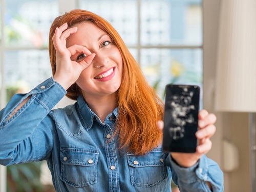 Mulher ruiva segurando smartphone quebrado com cara feliz sorrindo fazendo okey sinal com a mão no olho, procurando por entre os dedos
