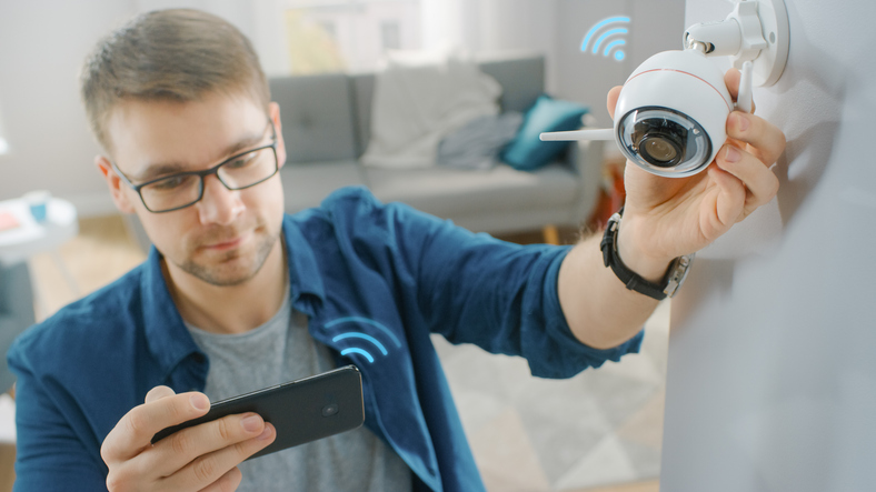 Jovem de óculos vestindo uma camisa azul está ajustando uma câmera de vigilância Wi-Fi moderna com duas antenas em uma parede branca em casa. Ele está verificando o feed de vídeo em seu smartphone.