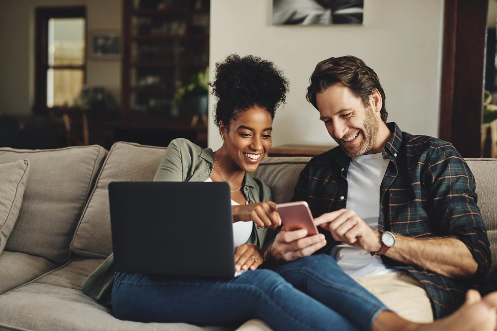 Casal feliz aprendendo a lucrar com a venda de celular usado no programa Trocafone, usando laptop e smartphone na sala de estar.