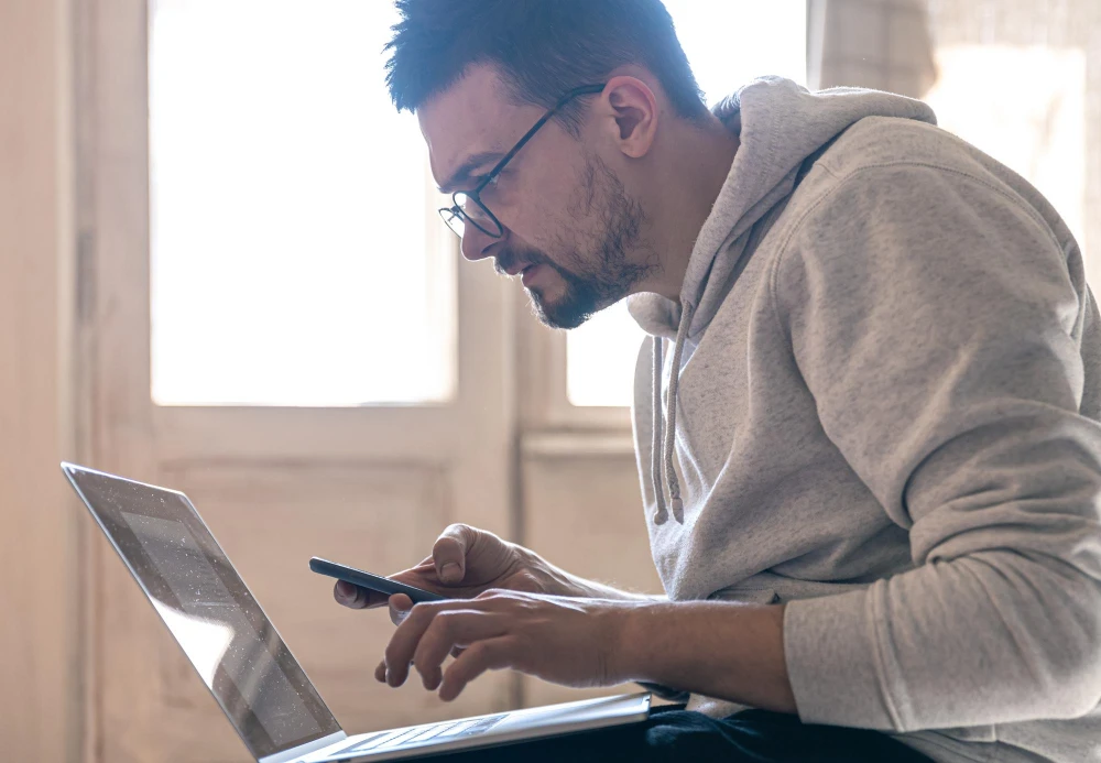 Homem com barba e óculos usando laptop e smartphone, sentado em ambiente interno bem iluminado. Conteúdo sobre como bloquear iphone roubado e icloud login.