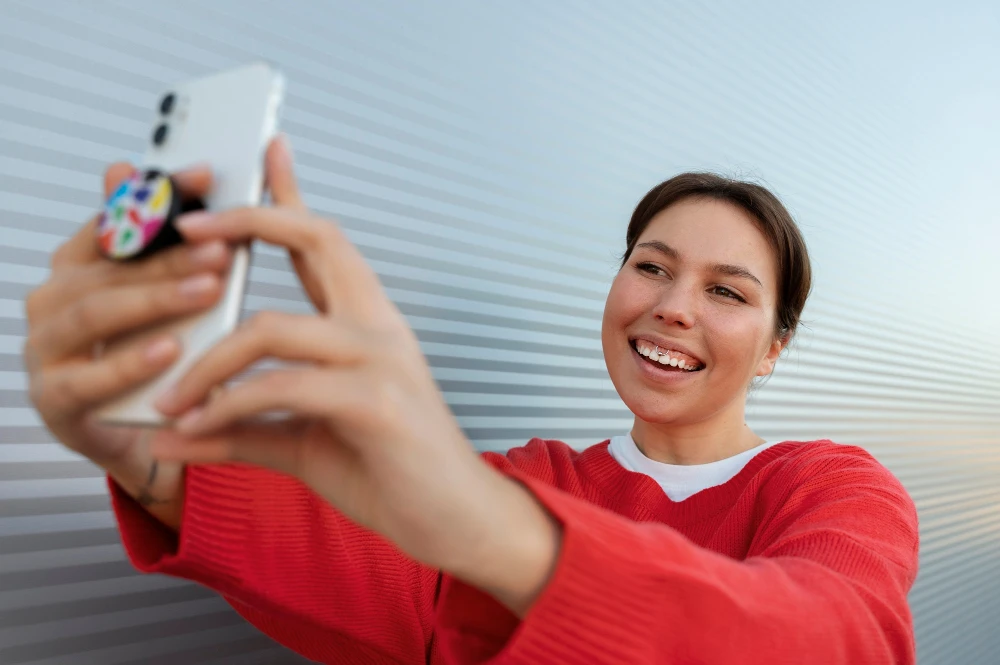 Jovem sorridente tirando selfie com smartphone na frente de uma parede moderna, usando blusa vermelha e acessório na mão, em ambiente bem iluminado.