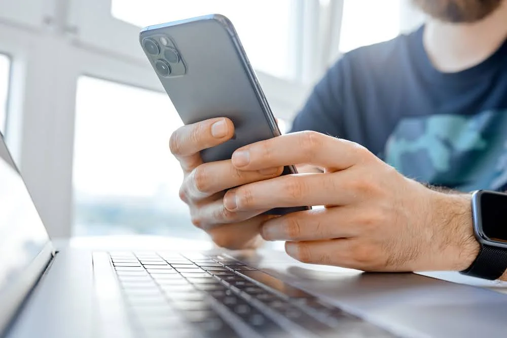 Pessoa usando smartphone cinza perto de laptop prata em mesa branca para conteúdo sobre "quanto vale meu celular usado?"