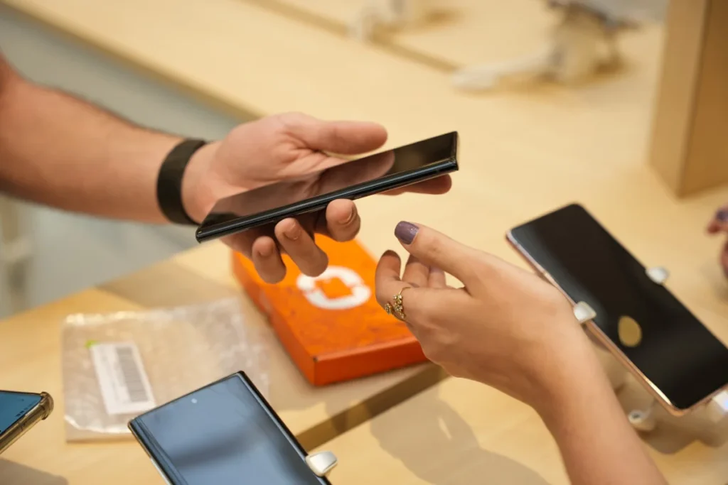 Pessoa segurando smartphone preto em loja de eletrônicos, com outra mão feminina próxima e celulares expostos na mesa, pra conteúdo sobre melhores celulares 2025