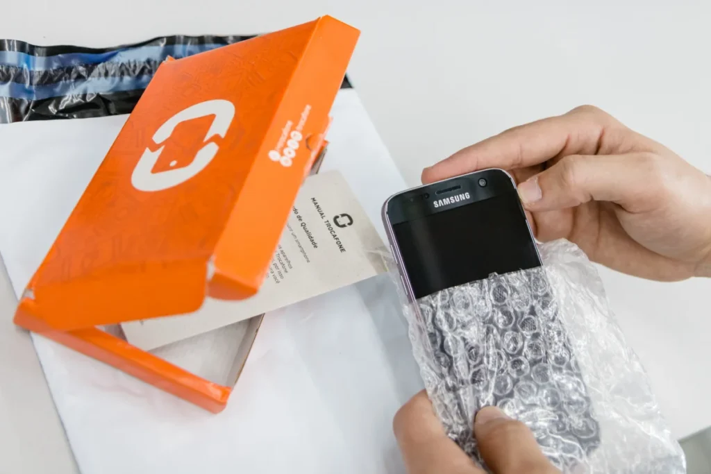 Pessoa desembrulhando celular Samsung preto de plástico bolha com embalagem laranja e manual para conteúdo sobre: Trocafone: compra ou venda seu celular sem sair de casa