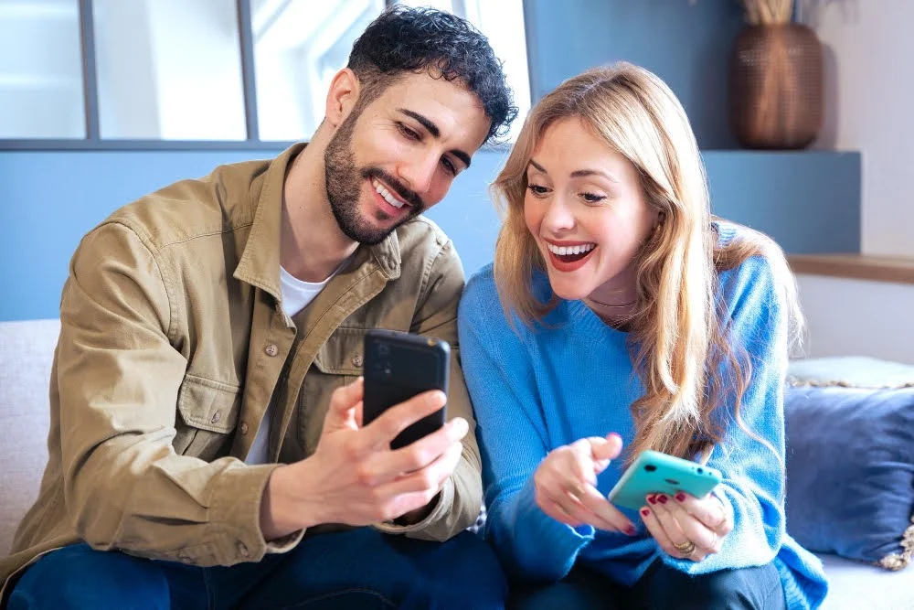 Casal sorridente vendo como comprar celular usado sentados em sofá em ambiente doméstico