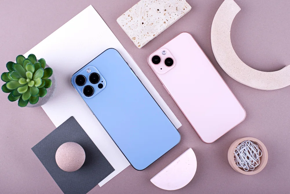 Dois smartphones, um azul e um rosa, dispostos sobre uma mesa com objetos decorativos minimalistas, incluindo uma planta suculenta, uma lua decorativa, e figuras geométricas em tons suaves. Conteúdo sobre iPhone 11.