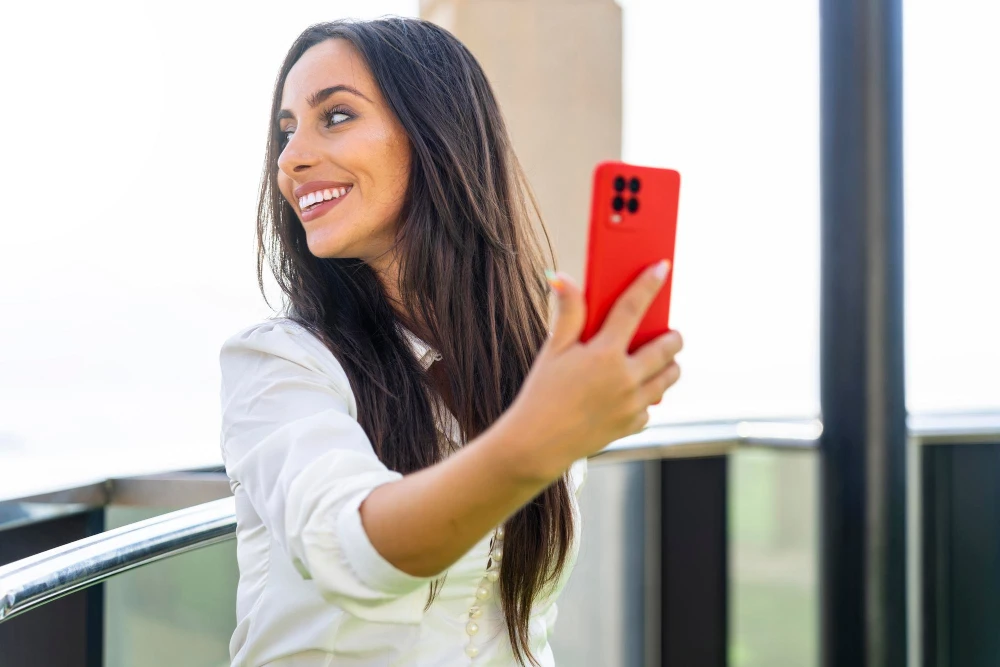 Mulher sorridente usando smartphone vermelho ao ar livre com fundo claro, destacando o melhor celular Motorola para fotos e entretenimento.