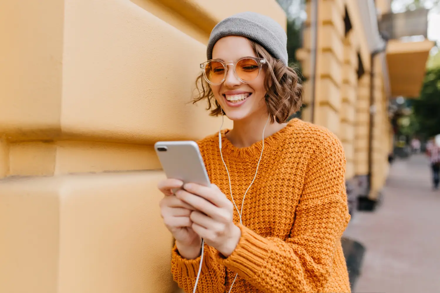 Jovem sorrindo ao usar o celular com fones de ouvido na rua, usando a tecnologia para como rastrear um celular por localização, de forma prática no dia a dia.