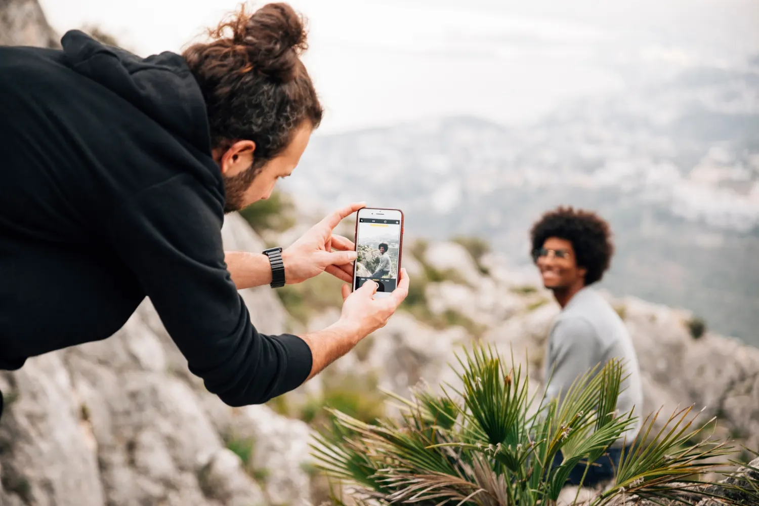 Homem em primeiro plano fotografa ao ar livre com um celular, usando a câmera para registrar uma pessoa ao fundo em cenário natural. Boa cena para destacar o melhor celular para fotos